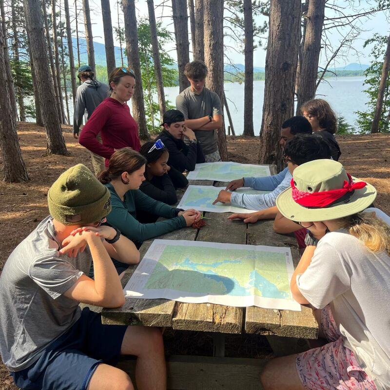 A group of people are gathered around a wooden table outdoors, looking at maps. They appear to be in a wooded area, possibly a park or campground, with a lake visible in the background. The group seems to be engaged in a map-reading activity, perhaps planning a hike or discussing the local geography. The atmosphere appears relaxed and collaborative.